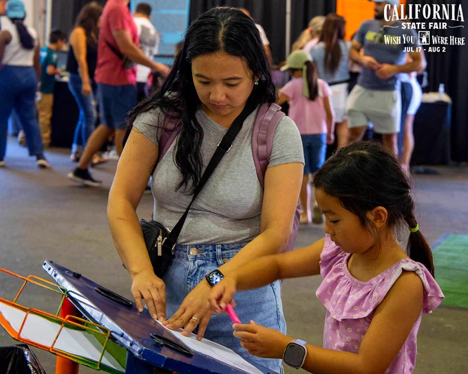 A woman and a young girl create a drawing at the California State Fair, surrounded by other visitors indoors.
