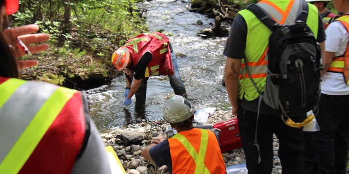 A group of individuals wearing high-visibility vests and helmets is gathered by a stream in a forested area. One individual is collecting a water sample from the stream. Others are observing or handling equipment on the rocky bank.