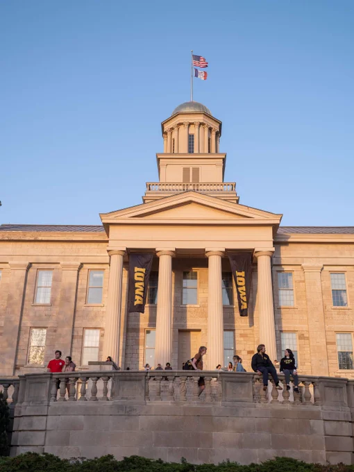 Old Capitol with Iowa banners under a clear blue sky, University of Iowa students stand in the front 