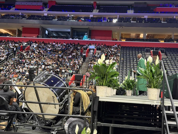 A wide shot of a graduation ceremony inside a massive, tiered arena filled with a large crowd. On the right, the lieutenant governor stands in a green graduation gown at a podium on a raised stage flanked by potted white peace lily plants. In the lower left foreground, members of a band sit behind a large Ludwig bass drum.