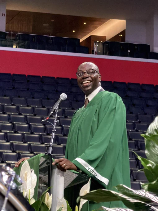 A close-up shot of the lieutenant governor smiling broadly at a podium during a graduation ceremony inside a large arena. He wears a green graduation gown with white trim that reads "TECH" on the sleeve over a white collared shirt and tie. He is looking off to the side, and white peace lily plants are visible in the foreground.