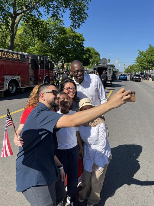 A group selfie taken during an outdoor street parade. A man in sunglasses and a dark blue shirt holds up a smartphone to take the photo. Smiling in the group is the lieutenant governor wearing a white shirt, a young boy, a woman with glasses, and a woman wearing a white hijab and sun hat. A red fire truck with "DEARBORN" printed on it is parked in the background under a clear blue sky.