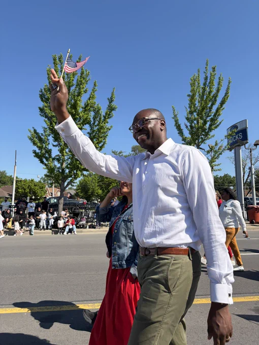 The lieutenant governor smiles while walking down a street during a sunny outdoor parade. He is wearing a white long-sleeve button-down shirt, olive green trousers, and a brown leather belt. He holds a small American flag high in his right hand. A woman in a denim jacket and red dress walks beside him, with spectators lined up along the sidewalk in the background.