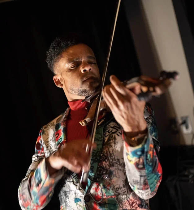 Musician wearing a colorful patterned jacket playing a violin with focused hand movements against a dark background.