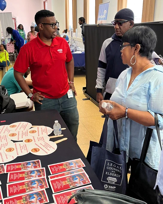 OIG staffer in a red shirt speaking with members of the community in front of a table with information about the agency.