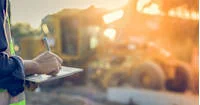 A person is writing on a clipboard at a construction site. In the background, there is heavy machinery, and the scene is bathed in warm sunlight.