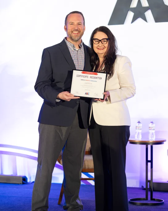 Two individuals are standing on a stage, smiling and holding a certificate of recognition. Behind them is a large logo featuring the letters "ASC." A small table with two water bottles is visible nearby.