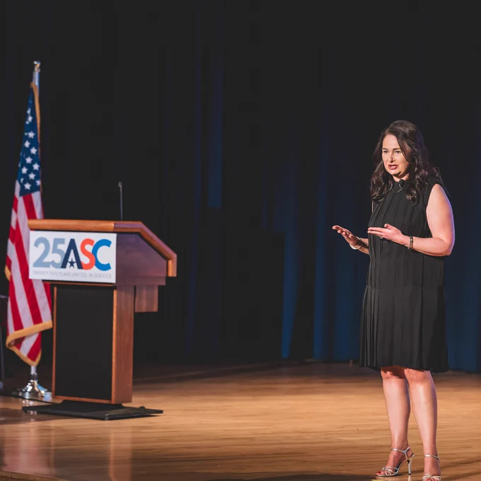 A person stands on a stage giving a speech, with a podium displaying the ASC logo nearby. An American flag is positioned next to the podium, and a banner reads "Uniting States in Service" and "StateCommissions.org.