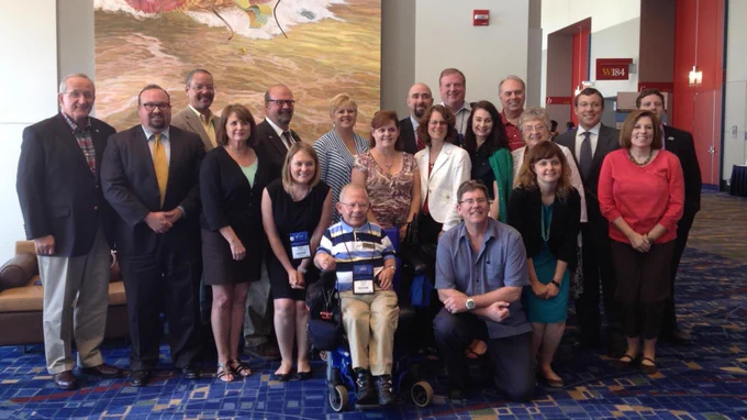 A group of people pose together in a spacious hallway with a mural depicting the Lewis and Clark Expedition in the background. There are various individuals standing and one seated in a wheelchair at the front. The setting appears to be a conference or event venue.