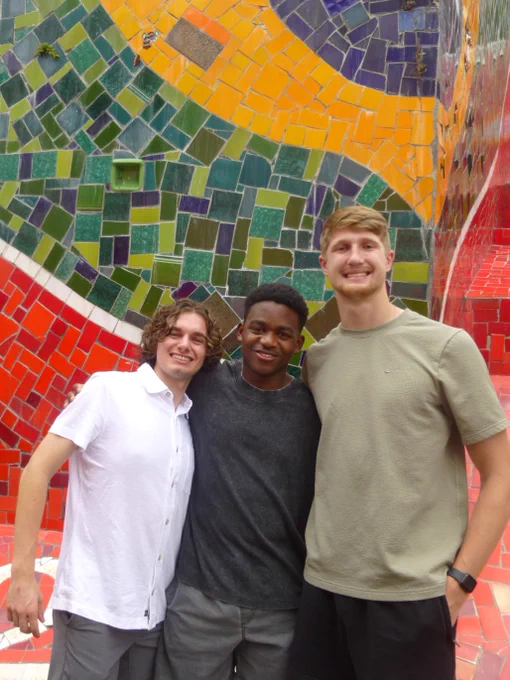 Three individuals smiling in front of a vibrant, colorful mosaic wall.