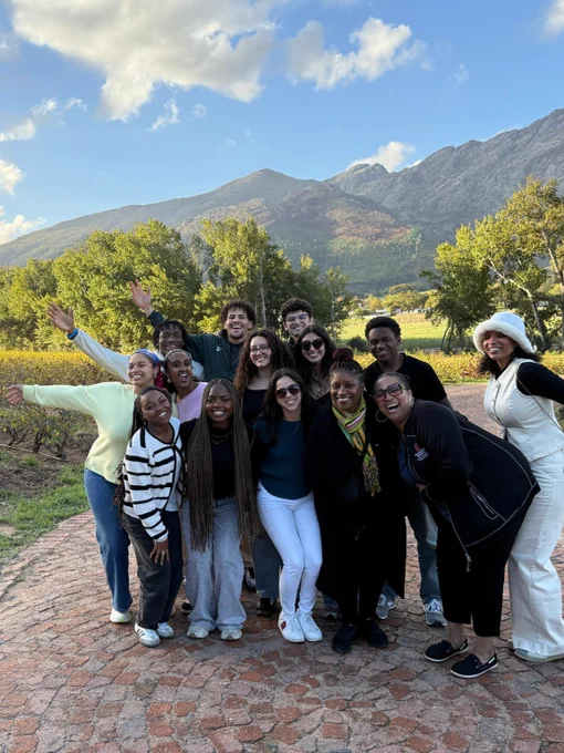 A group of people smiling and posing outdoors with a background of scenic mountains and trees. The sky is clear with some clouds, and the ground is paved with stones. Everyone appears cheerful and is dressed casually.