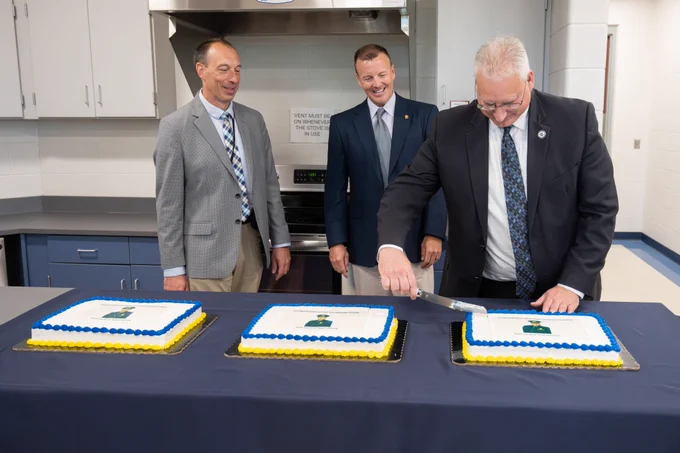 two men stand beside each other while another cuts cake sitting on table