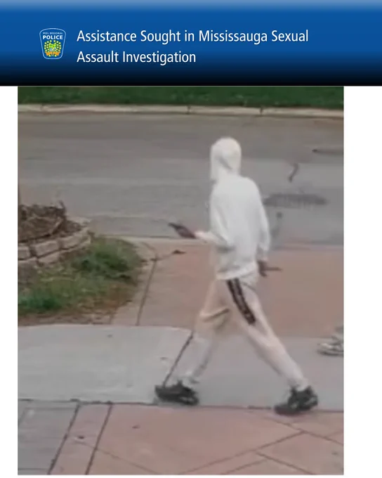 Person in white hoodie and light pants walking on sidewalk near street, holding a phone in one hand.