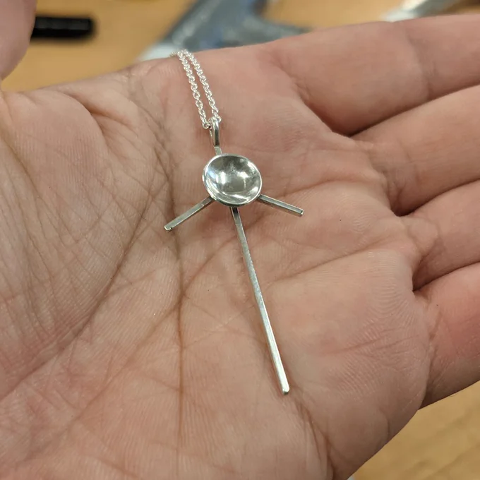 A silver Voyager inspired pendant shaped with a domed dish with radiating antenna rods on a fine chain, held in an open hand.
