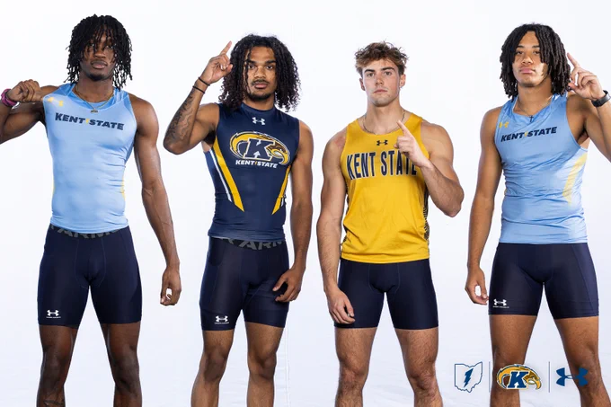 Four athletes from Kent State University, wearing athletic uniforms, stand together against a plain background. Two individuals are in light blue uniforms, one is in a navy and gold uniform with "KS" insignia, and another is in a gold top with "Kent State" written across. They are posing with one finger raised. The Kent State and Under Armour logos are displayed below.