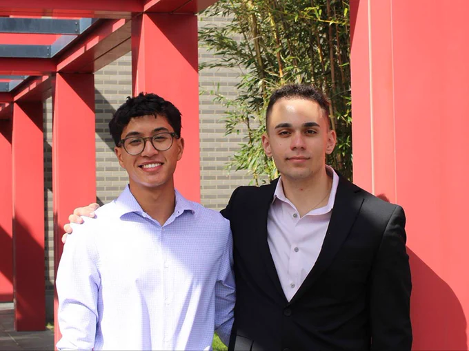 Two people are standing together side by side, smiling at the camera. They are in an outdoor setting with red pillars and greenery visible in the background. One person is wearing glasses and a light blue shirt, and the other is in a black suit.