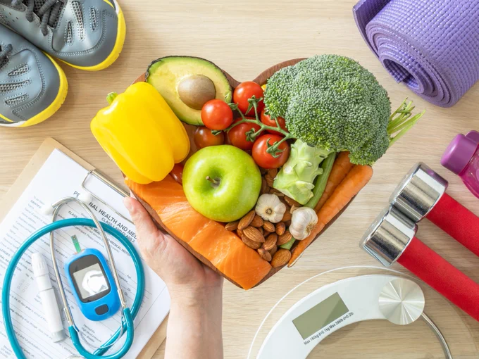A hand holds a heart-shaped wooden bowl filled with colorful vegetables and fruits, including broccoli, an avocado, cherry tomatoes, a yellow bell pepper, apple slices, almonds, and garlic. Surrounding items include a stethoscope, a glucometer on a medical form, running shoes, a rolled yoga mat, dumbbells, and a digital bathroom scale.