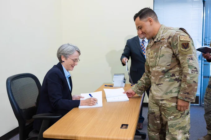 A person in military uniform is handing a copy of "Get Back Up: Lessons in Servant Leadership" to UTEP President Heather Wilson who is seated and signing paperwork at a wooden table. The seated person is smiling. In the background, a person in a suit is holding a book and a pen.