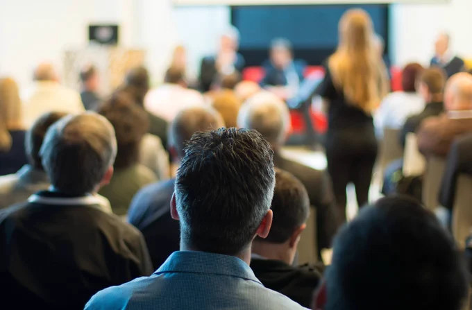 A group of people seated in a conference room, facing a panel of speakers. The focus is on the audience from behind, with blurred figures of the panelists and a presenter in the background.