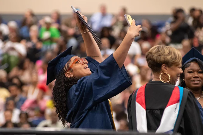 A group of graduates in academic regalia, one person raising their arms in celebration, surrounded by a crowd.