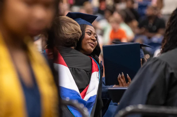 A graduation ceremony with a woman in a cap and gown smiling and embracing another person in the crowd.