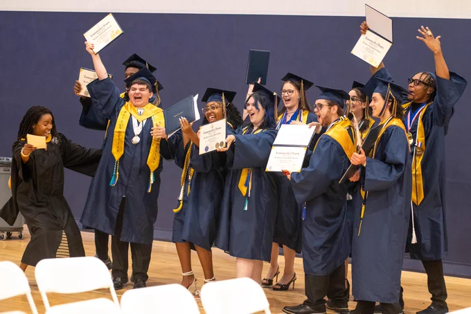 A group of graduates in blue and gold gowns celebrating their achievement, holding diplomas and raising their arms in the air.