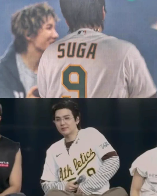 SUGA from BTS wears a white Athletics jersey with green and gold lettering. The back of the jersey reads, “SUGA” with the number 9.