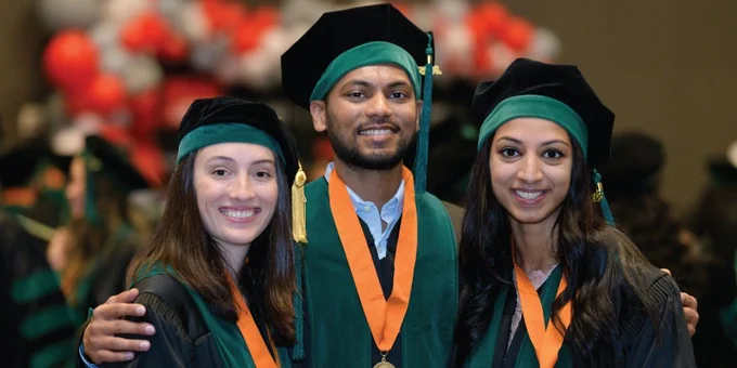 Three individuals in graduation attire stand closely together, smiling at the camera. They are wearing black caps and green gowns with orange stoles. In the background, a collection of red and white balloons is visible.