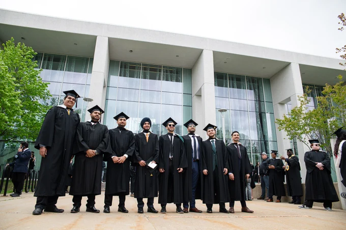 A group of graduates in caps and gowns stand in front of a modern building with large windows. The building is surrounded by trees.