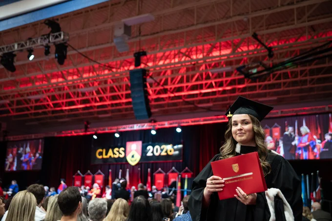A graduate in a cap and gown holds a red degree cover, standing among an audience at an indoor graduation ceremony. The backdrop shows a stage with the words "Class of 2026" mounted. Red lighting highlights the ceiling and stage area.