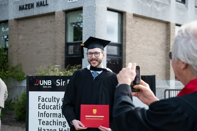 A graduate is smiling and holding a degree in front of Hazen Hall at the UNB Saint John campus. Another person is taking a photo of the graduate.