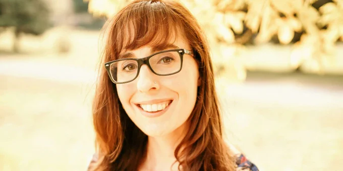 Person with glasses and long hair wearing a floral patterned blouse standing outdoors under a tree with golden leaves.