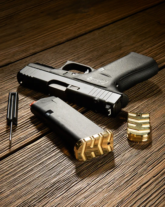 Compact pistol with a magazine fitted with a machined brass magazine base plate resting on a wooden surface. A second brass base plate and installation tool are displayed nearby.