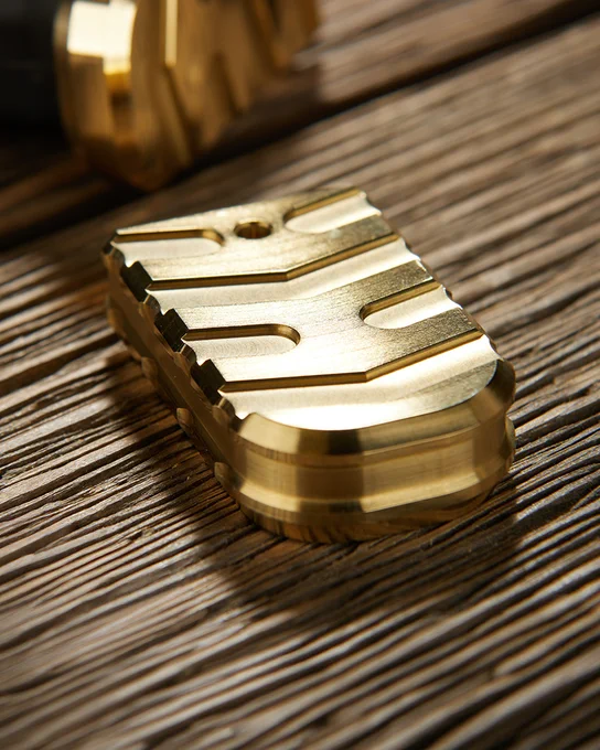 Detailed close-up of a solid brass magazine base plate featuring precision CNC machining, textured grip cuts, and a polished metallic finish on a rustic wood background.