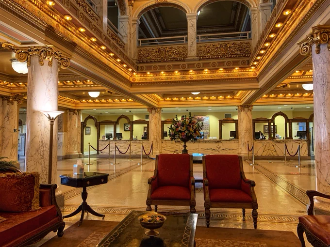 French Lick Springs Hotel lobby featuring grand marble columns and an intricately decorated ceiling. The space is elegantly lit with warm lighting. In the foreground, there are plush red chairs and decorative tables. In the background, the reception desk is visible with framed mirrors and floral arrangements enhancing the luxurious atmosphere.