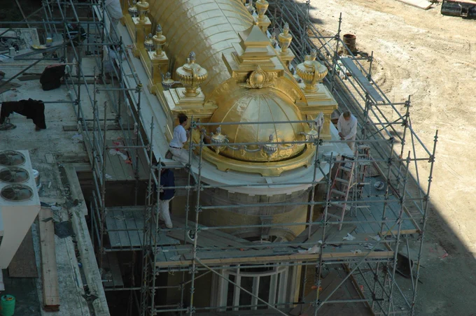 The image shows a group of workers on scaffolding performing restoration work on the golden dome of 1875: The Steakhouse at French Lick Springs Hotel. The dome is adorned with ornate decorations, and the scaffolding surrounds it to provide access for maintenance.