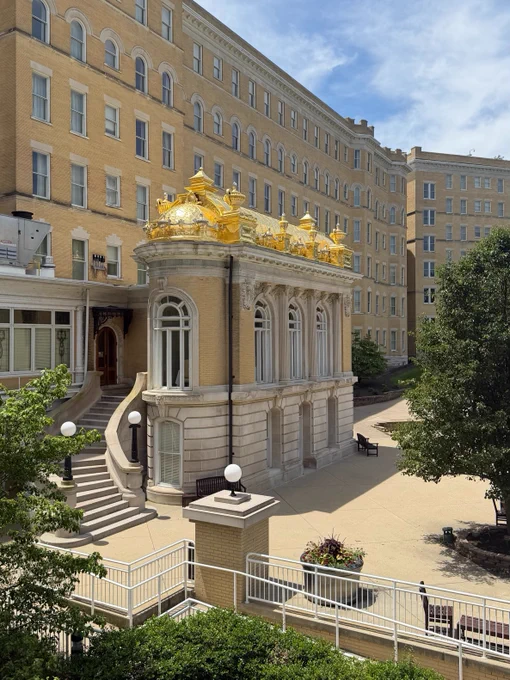 A historic French Lick Springs Hotel with a light brick exterior featuring an ornate golden roof structure. The setting includes a courtyard with trees, plants, and a staircase leading to an arched entrance. The sky is partly cloudy with sunlit areas.