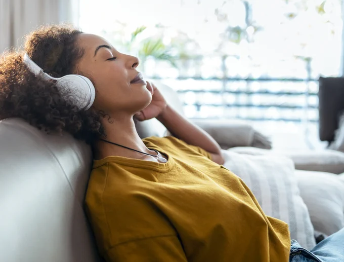 A person wearing headphones relaxes on a sofa with eyes closed, appearing to enjoy music. Natural light filters through a window, illuminating the cozy setting.