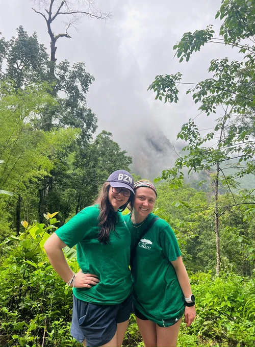 Two people in green shirts stand closely together, smiling in a lush, green forest. They are wearing caps and appear happy, with misty mountains visible in the background.