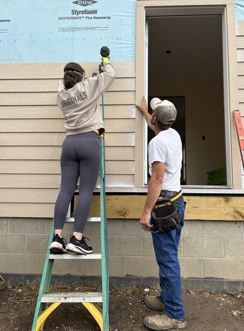 Two people work on the exterior of a building. One stands on a ladder drilling into wooden siding, while the other holds a board. 