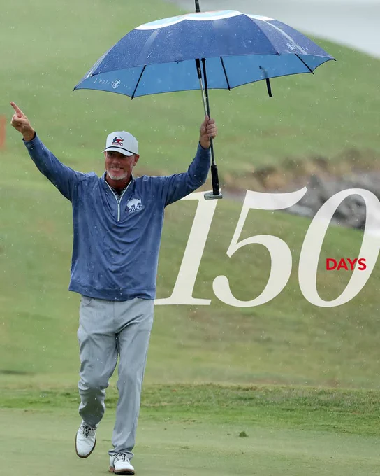 A person walking on a golf course, holding a large blue and white golf umbrella. The individual is wearing a cap, long-sleeved jacket, and pants. They have one arm raised in the air. Large numbers "150 Days" are overlaid on the image.