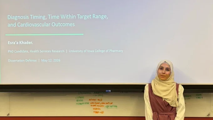 A woman named Esra’a Khader wearing a light-colored hijab and a mauve dress stands beside a presentation slide titled “Understanding Hypertension in Young Adults.” The slide includes details about diagnosis timing, cardiovascular outcomes, and a University of Iowa College of Pharmacy dissertation defense presentation.