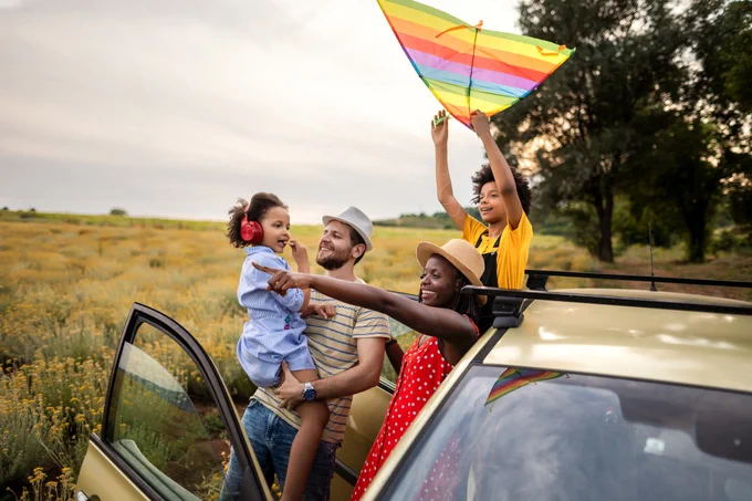 A group of people enjoying a day outdoors next to a car. One person is holding a child, who has headphones on. Another person is raising a colorful kite. The setting is a field with trees and a cloudy sky.