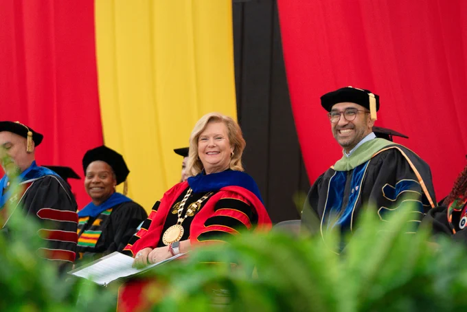 A group of faculty members in academic regalia, with a woman wearing a gold medallion and a man beside her smiling. Red and yellow drapes hang in the background.