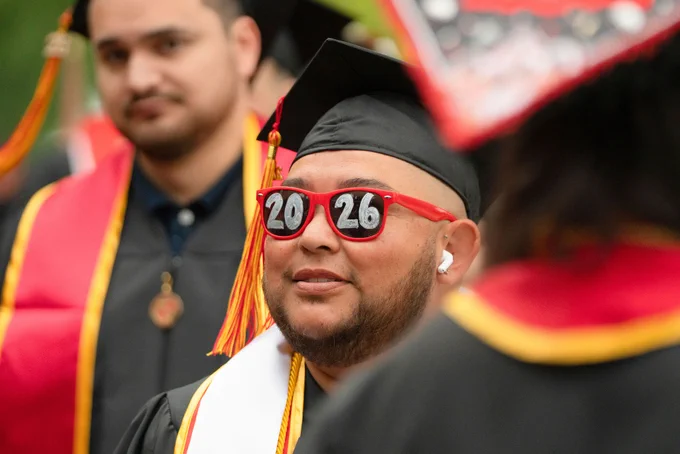 Graduate with sunglasses showing "2026," smiling during a ceremony. Wears cap and gown with red and yellow accents, conveying joy and achievement.