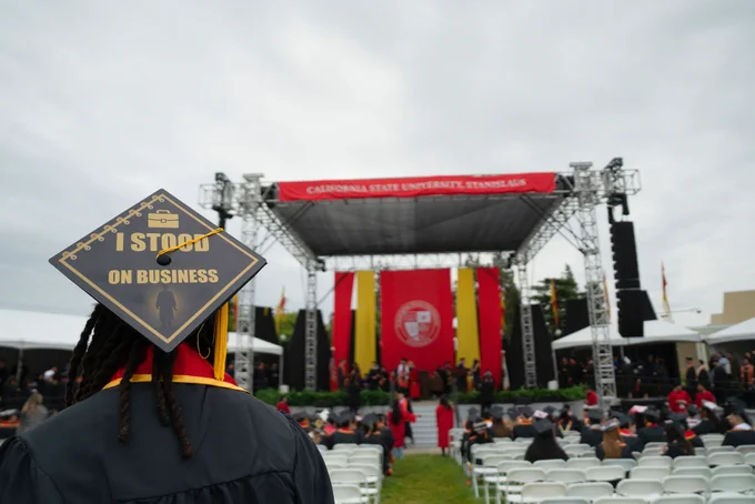A graduate stands facing an outdoor stage at California State University, Stanislaus. Their cap reads "I STOOD ON BUSINESS," conveying pride and achievement.