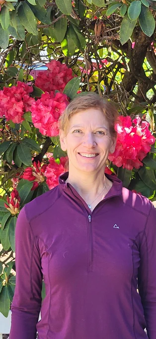 Portrait of Gail Hering smiling in front of a blooming rhododendron with bright pink flowers. She is wearing a long-sleeve purple quarter-zip top and standing outdoors in natural sunlight.