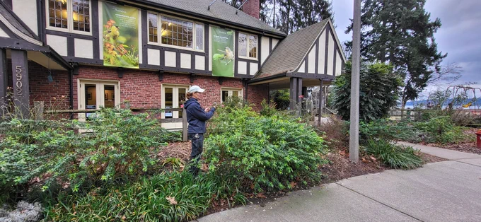 Gail Hering stands in a landscaped garden outside a brick-and-timber building, observing plants near a pathway. Trees, shrubs, and garden beds surround the area, with a playground visible in the background.