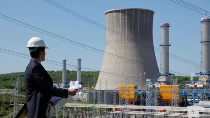 A person wearing a hard hat is holding papers and observing a power plant with a prominent cooling tower and multiple smokestacks. Transmission lines are visible overhead, and there is greenery in the background.