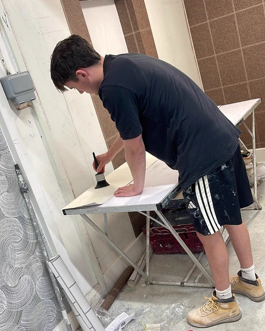 Person wearing a black shirt and shorts applying paint with a brush on a white surface in a workshop setting.